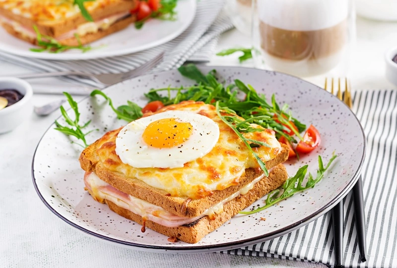 Close-up of a Croque Madame sandwich topped with a sunny-side-up egg, served with arugula and cherry tomatoes on a speckled plate.