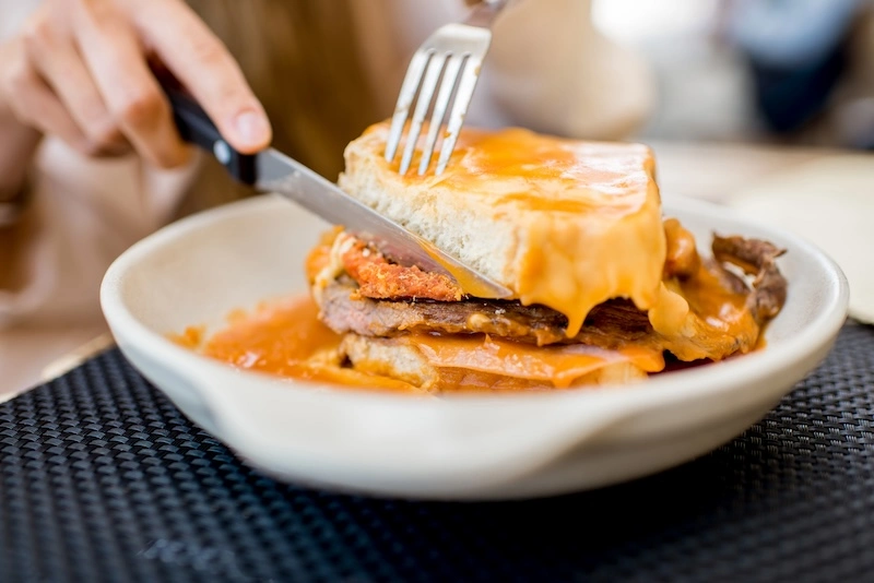 Portuguese Francesinha sandwich being cut, covered in melted cheese and drenched in rich tomato-beer sauce.