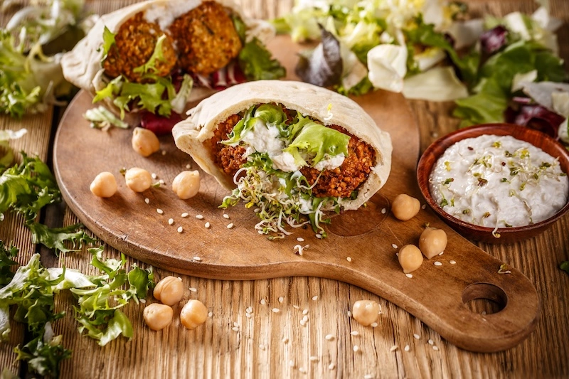 A falafel pita sandwich stuffed with crispy chickpea balls, shredded lettuce, sprouts, and creamy tahini sauce, resting on a wooden board with scattered chickpeas and greens.