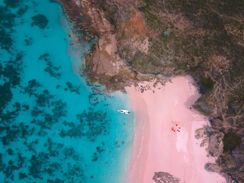 Aerial view of a boat anchored near a stunning pink sand beach with turquoise waters and rocky shoreline on Komodo Island, Indonesia.