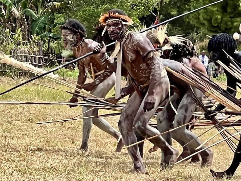 Indigenous men in Papua New Guinea wearing traditional body paint and headdresses perform a ceremonial dance while holding spears in an open grassy area.