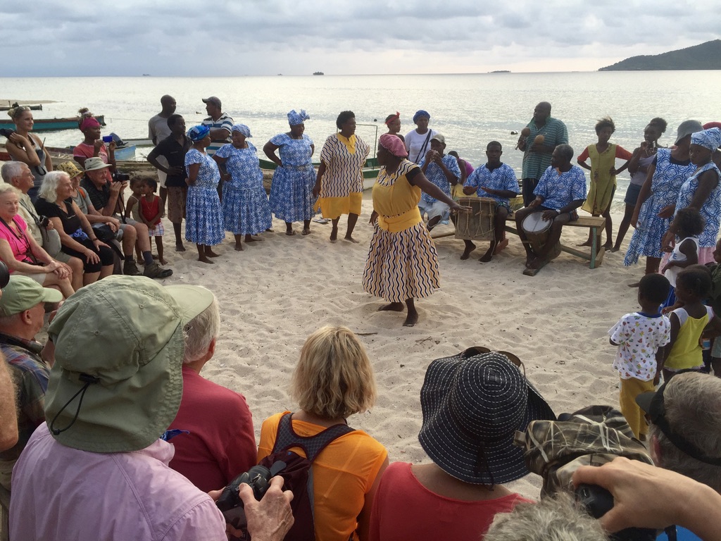 Local community members perform a traditional dance on a beach while a small group of travelers watches and engages.