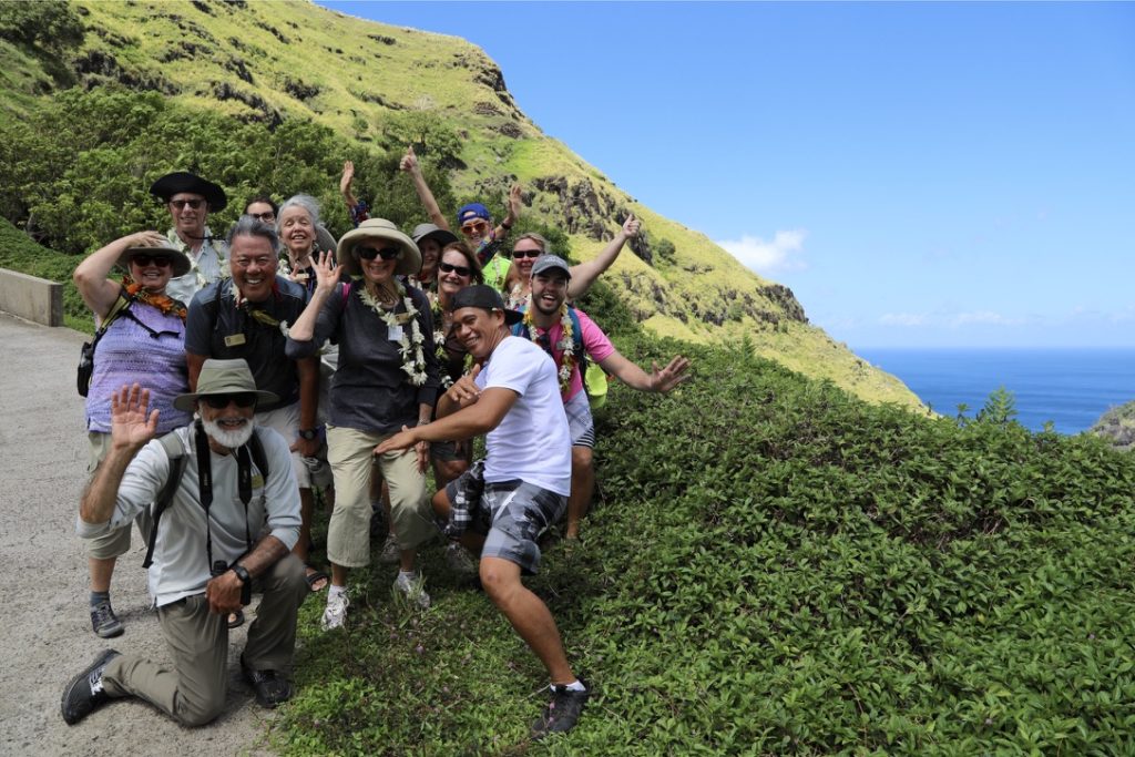 A small group of travelers hikes along a green coastal trail with a local guide, smiling and interacting as they walk.