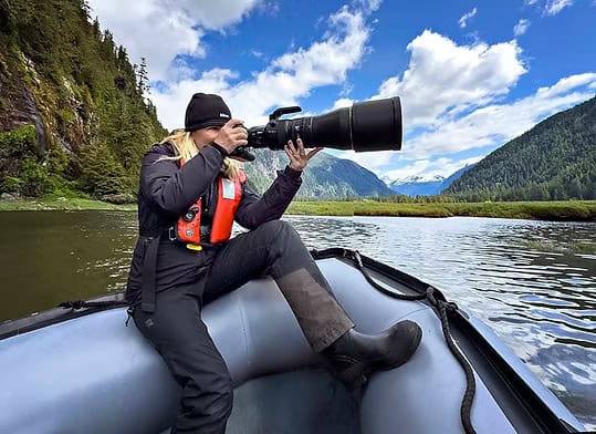 Michelle Valberg photographing wildlife from a boat on a river surrounded by mountains