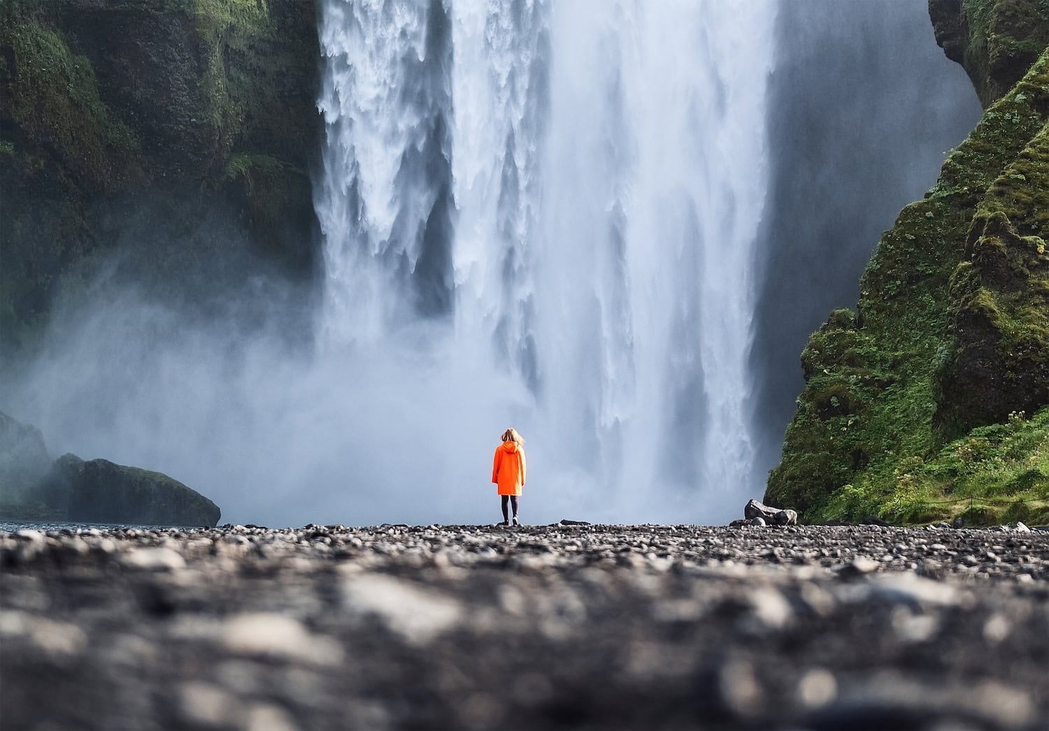 Traveler in orange rain jacket standing before the powerful cascade of Skógafoss waterfall on Iceland's South Coast